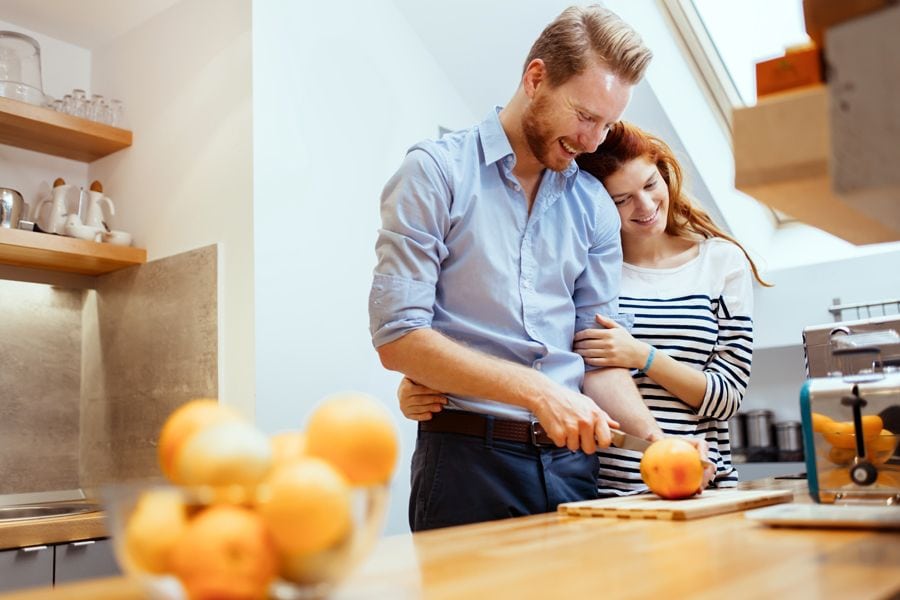 Three Reasons to Consider a Geothermal Heat Pump. Image shows man and woman in kitchen. Man is cutting and orange on a small wooden cutting board and the woman is resting her head on his shoulder and smiling.